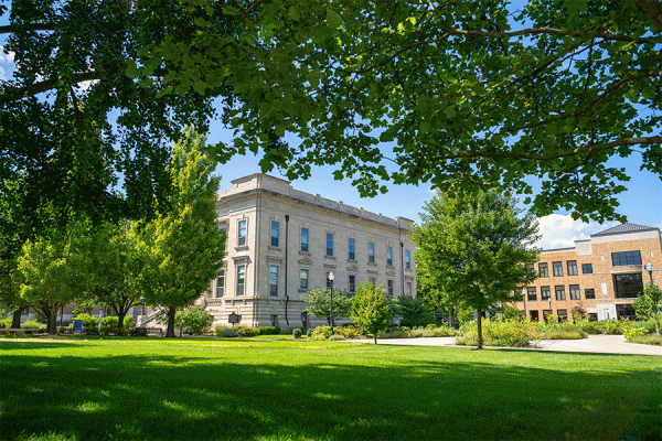 A grey stone building, Normal Hall, is seen at a distance from beneath a canopy of green trees. It is summer, the sun is out, and the grass is green. The reddish-brown brick Science Building is also visible to the left of Normal Hall.